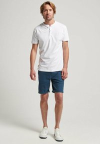 Camiseta henley de manga corta blanca, pantalones cortos azules marinos y zapatillas blancas. Conjunto casual con un ajuste relajado y elementos de diseño mínimos.