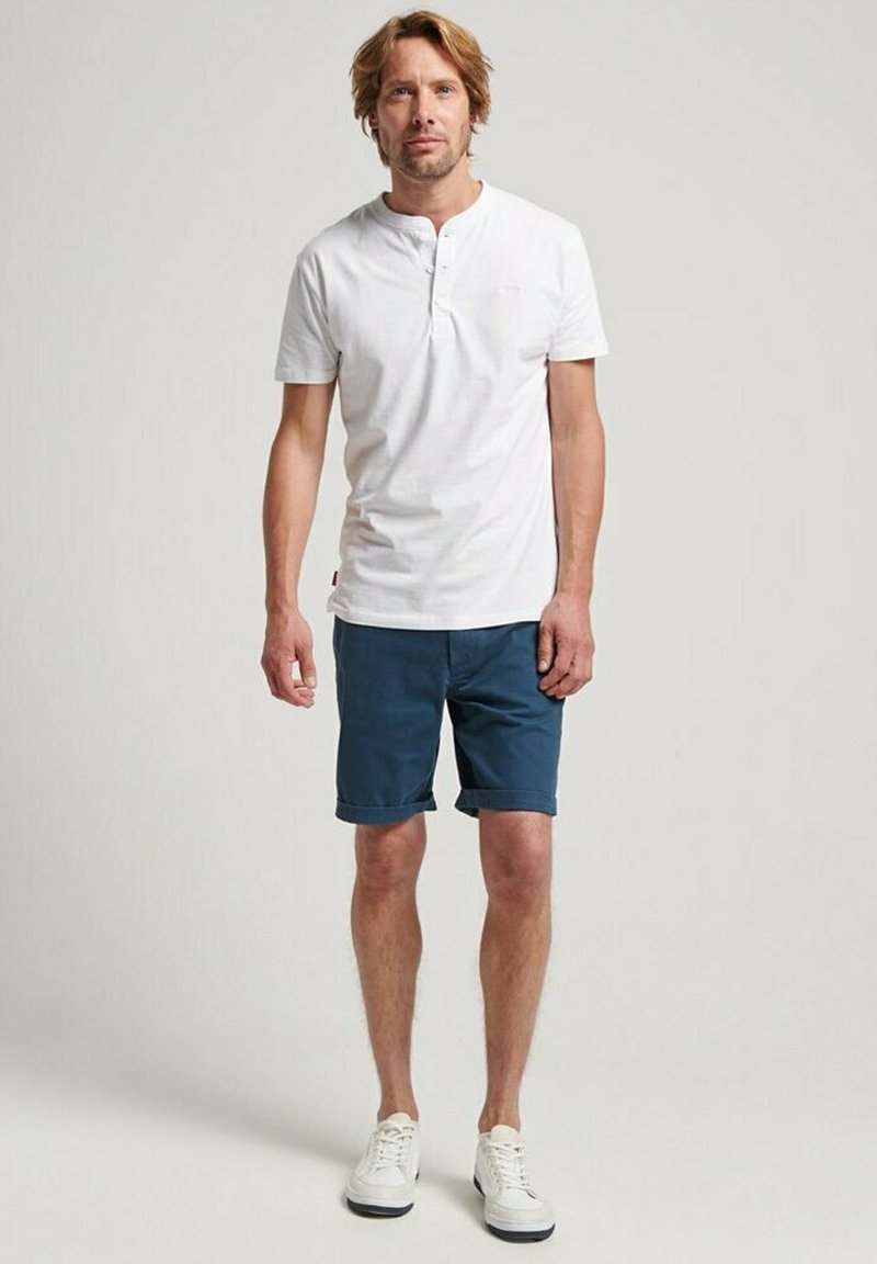 Camiseta henley de manga corta blanca, pantalones cortos azules marinos y zapatillas blancas. Conjunto casual con un ajuste relajado y elementos de diseño mínimos.