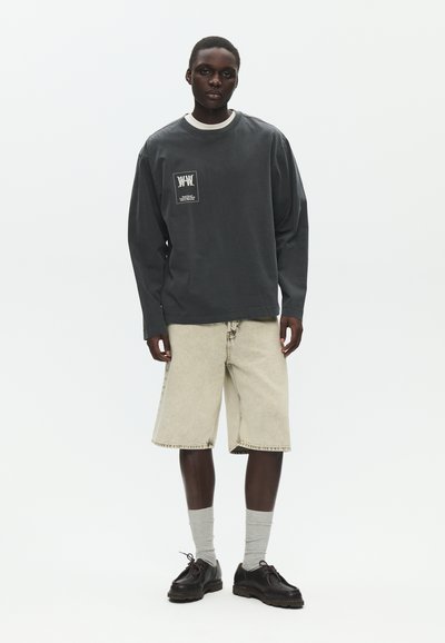 Jeune homme portant une chemise à manches longues sombre avec un écusson, un short beige clair jusqu'aux genoux, des chaussettes grises et des chaussures noires à lacets, debout devant un fond blanc.