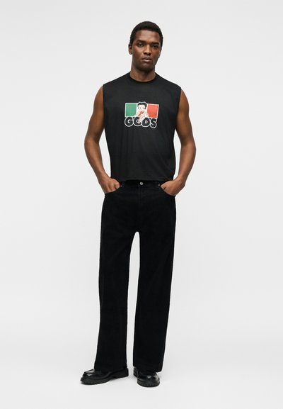 Άνδρας στέκεται με τα χέρια στις τσέπες, φοράει μαύρο αμάνικο πουκάμισο με κόκκινο, λευκό και πράσινο σχέδιο και μαύρο παντελόνι με φαρδιά μπατζάκια και μαύρα παπούτσια.