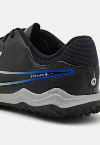 Zapatilla deportiva negra y gris con una franja azul y plateada, texto "TIEMPO", suela texturizada y un cuello de tela acolchado, mostrada desde un ángulo posterior.