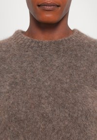 Nahaufnahme einer Person, die einen samtigen braunen Rundhalsausschnitt-Sweater trägt, wobei das untere Gesicht, der Hals und die obere Brust zu sehen sind.