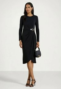 Sieviete, tērpta melnā midi kleitā ar garām piedurknēm un sānu sprādzi, tur melnu rokassomu un valkā melnas augstpapēžu sandaļus.