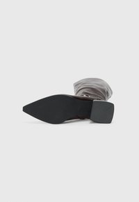 Bout en pointe, semelle noire et talon carré d'une botte en cuir marron avec une partie supérieure souple plissée sur un fond blanc.