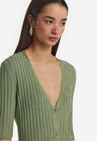 Stickad grön cardigan med djup V-ringning och korta ärmar, med vertikal ribbad textur och dekorativa knappar.