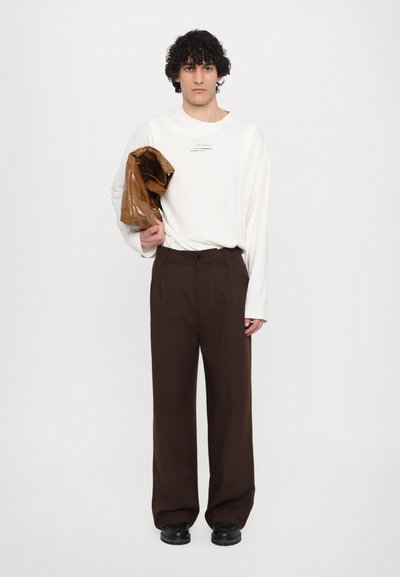 White long-sleeve shirt with a label, brown wide-leg trousers, holding a shiny brown jacket, and black footwear.