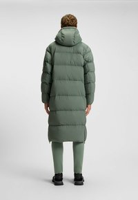 Leichter, gepolsterter langer Mantel in Hellgrün mit Kapuze, horizontal gesteppten Abschnitten, Frontreißverschlüssen und bis zur Wade reichend.