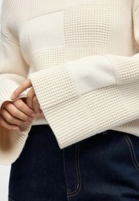 Jersey crema con patrones texturizados y acanalados, mangas anchas y un corte holgado, combinado con tejido de denim oscuro.