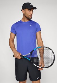 Άνδρας αθλητής φορώντας μπλε κοντομάνικο μπλουζάκι της Nike και μαύρες βερμούδες, κρατώντας ένα τένις ρακέτα με μπλε λεπτομέρεια. Μαύρο καπέλο στο κεφάλι.
