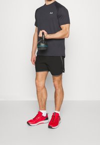 Pantalones cortos deportivos negros con detalles grises, una camiseta de manga corta gris oscuro y zapatillas rojas. Sosteniendo un kettlebell negro con detalle azul.
