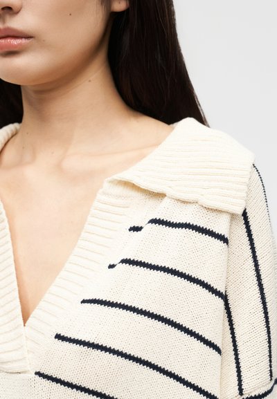 Γυναίκα που φοράει εκρού πλεκτό πουλόβερ με μαύρες οριζόντιες ρίγες και φαρδύ γιακά, που αφήνει εμφανή τον λαιμό και το πάνω μέρος του στήθους.