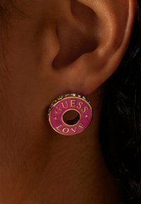 Oorring in goudkleurig metaal met een roze geëmailleerd oppervlak waarop de tekst "GUESS LOVE" staat, omringd door kleine heldere kristallen en een centrale opening.