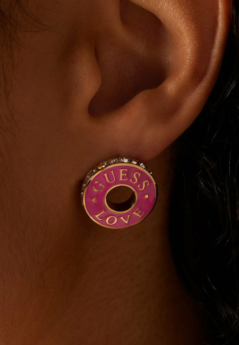 Oorring in goudkleurig metaal met een roze geëmailleerd oppervlak waarop de tekst "GUESS LOVE" staat, omringd door kleine heldere kristallen en een centrale opening.