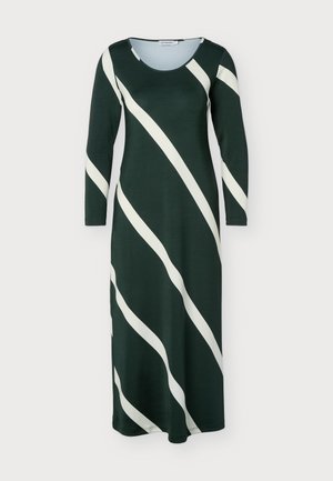 Femme portant une robe midi à manches longues vert foncé avec de larges rayures diagonales beige, debout droite avec des talons noirs à bout pointu.