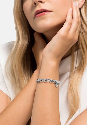 Zilveren armband met een schakelklinkontwerp en sprankelende accenten, versierd met een klein hartje, gedragen om een pols tegen een effen achtergrond.