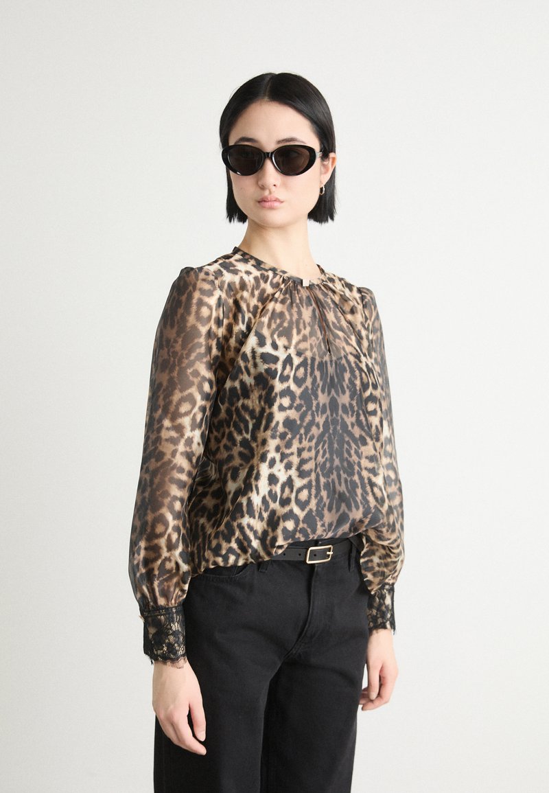 Blouse en imprimé léopard transparent avec encolure froncée et accents en dentelle noire au niveau des poignets, assortie à un pantalon noir et des lunettes de soleil noires en forme ovale.
