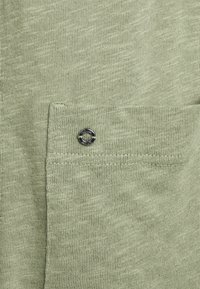 Tejido de punto verde claro con un pequeño bolsillo en el pecho. Presenta un detalle de botón circular plateado en el bolsillo. Apariencia suave y texturizada.