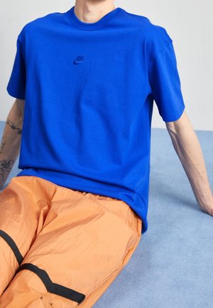 Osoba ubrana w jaskrawo niebieską koszulkę Nike i pomarańczowe spodnie z czarnymi paskami, siedząca na jasnoniebieskiej powierzchni.