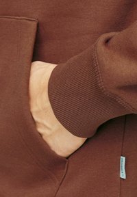 Barna pulóver anyag, texturált felülettel, bordás mandzsettával és elülső zsebbel, amelyhez egy diszkrét anyagcímke van hozzárendelve.