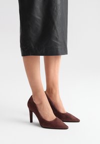 Bordowe zamszowe szpilki z noskiem w szpic i smukłym obcasem stiletto, zestawione z czarną skórzaną spódnicą do kolan.