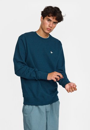 Jeune homme ajustant la manche d'un sweat-shirt bleu foncé avec un petit logo blanc de mouette, portant un pantalon bleu clair devant un fond uni.