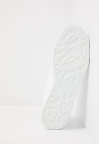 Vit sneakersula med abstrakta, vågiga mönster lutande mot en vit hörnvägg på en enfärgad vit yta.