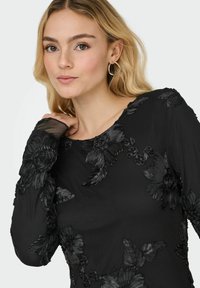 Czarny top z długim rękawem, ozdobiony teksturowanymi aplikacjami kwiatowymi i okrągłym dekoltem, wykonany z miękkiego, lekkiego materiału.