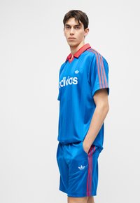 Νέος άνδρας στέκεται με τα χέρια στις τσέπες, φορώντας μπλε αθλητική ενδυμασία Adidas με κόκκινες ρίγες και γιακά, πάνω σε απλό φωτεινό φόντο.