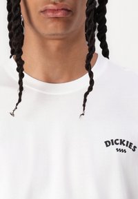 Fehér garbós póló, amelyen egy fekete "DICKIES" logó található a bal mellen. A szövet sima, az anyag textúrája jól látható.