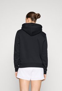 Hoodie preto feito de um tecido de algodão misto, apresentando um capuz grande e um acabamento canelado. Usado com calções brancos, visto de trás.