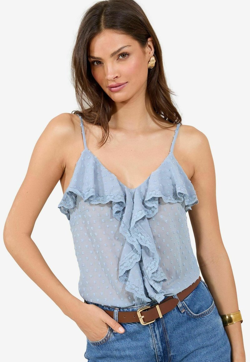 Blusa azzurra chiara, semi-trasparente, con dettagli in pizzo arricciato e texture a pois, abbinata a jeans a vita alta e una cintura marrone.