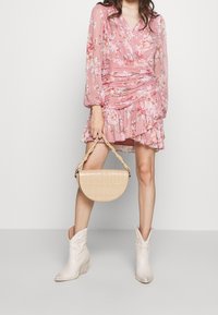 Kemeja wrap bunga merah jambu dengan lengan panjang, hem berkedut, dan beg tangan setengah bulan beremboss buaya berwarna beige; dipadankan dengan but koboi putih.