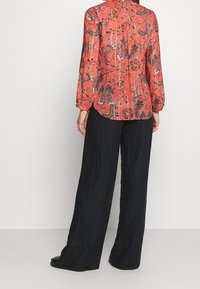 Blouse rouge à motifs floraux avec des manches longues et une emmanchure au dos. Associée à un pantalon noir ample. Le tissu texturé semble léger.