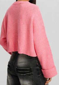 Femme portant un pull en tricot rose ample avec les poignets retroussés et un jean noir délavé, vue de dos sur un fond uni.