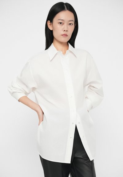 Biely košeľový vrch s golierom, dlhými rukávmi a voľným strihom; vyrobený z ľahkej látky s hladkou textúrou.