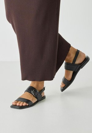 Czarne skórzane sandały na płaskiej podeszwie, z szerokimi paskami i detalem klamry, zestawione z prążkowanymi, ciemnobrązowymi spodniami.