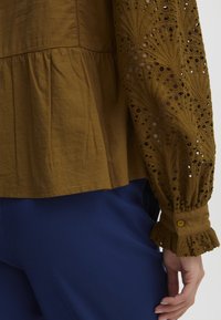 Top in cotone marrone con maniche lunghe testurizzate e ricamate, caratterizzato da modelli a taglio e da un polsino arricciato; indossato sopra dei pantaloni blu.