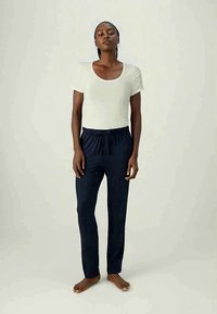 Woman with braided hair stands barefoot wearing a white short-sleeve top and dark drawstring pants on a plain background.