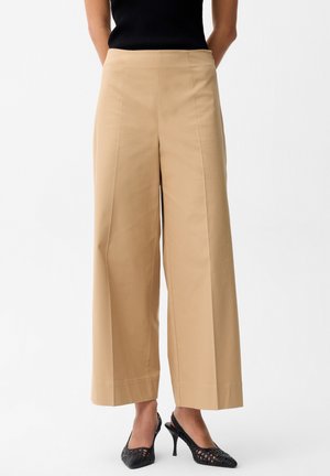 Kobieta ubrana w beżowe szerokie spodnie typu cropped oraz czarny bezrękawnik i czarne plecione buty na obcasie z ostrym noskiem.