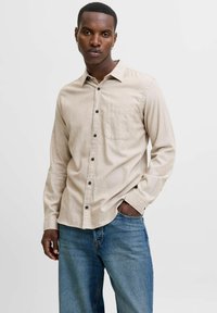 Man with short hair and earrings wearing beige button-up shirt and blue jeans standing with one hand in pocket against white background.