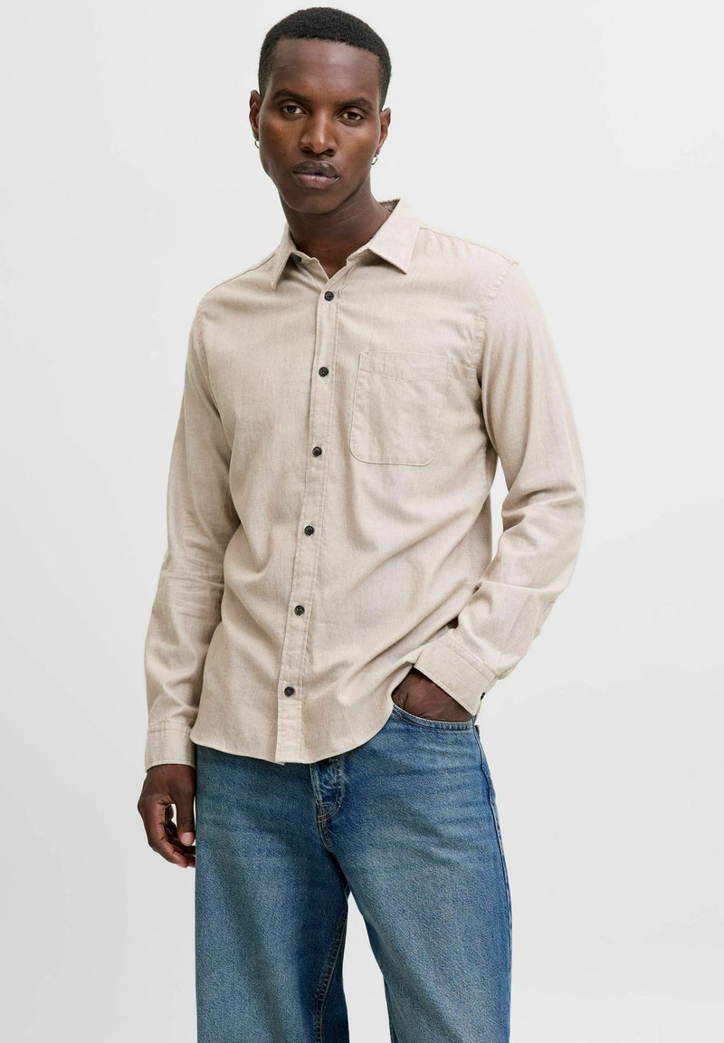 Man with short hair and earrings wearing beige button-up shirt and blue jeans standing with one hand in pocket against white background.