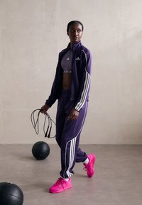 Violet sportinė apranga su baltais dryžiais, įskaitant užtraukiamą striukę ir atitinkančius kelnes. Ryškiai rožiniai sportbačiai ir šokdynė rankoje.