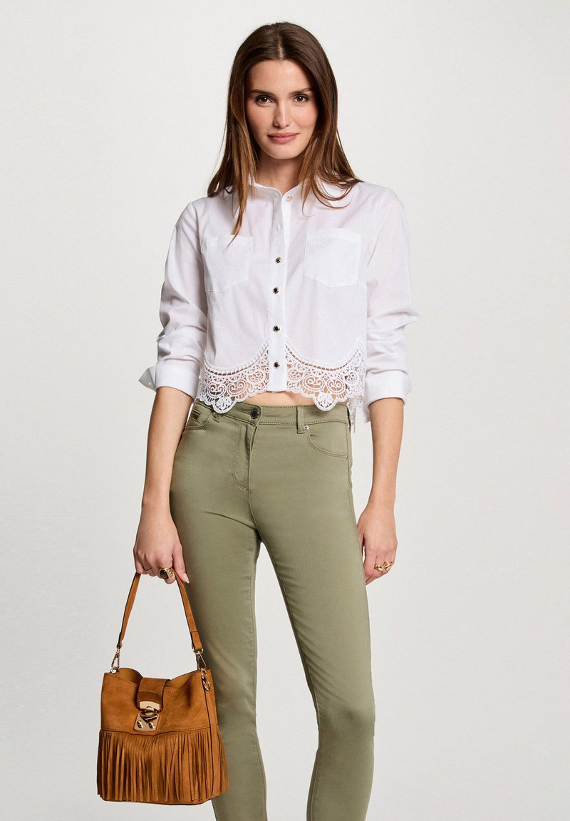 Weißes, kurzes Oberteil mit Spitzenbesatz, zwei Brusttaschen, kombiniert mit grünen Skinny-Jeans. Hält eine braune Wildledertasche mit Fransen und einem Verschluss.