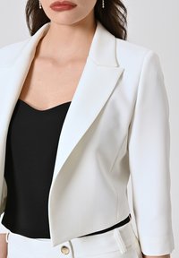Blazer blanc sur mesure avec un col cranté, des épaules structurées et des manches trois-quarts, associé à un haut noir à encolure cœur.