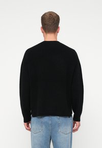 Suéter negro de punto con cuello redondo y mangas raglán. Presenta una textura de canalé y un corte relajado. Combinado con jeans de mezclilla en un tono azul claro.