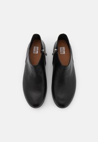 Zapatos de piel negra tipo slip-on con textura suave, punta redondeada y cremalleras a los lados. El interior presenta un forro de piel marrón.