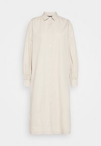 Longue robe beige à boutons avec manches longues bouffantes, col pointu et poignets ajustés sur un fond uni.