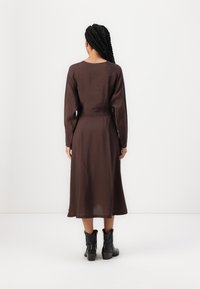 Brun, långärmad klänning med en åtsittande midja och utsvängd kjol. Tillverkad av ett slätt tyg, bärs med svarta ankelstövlar.