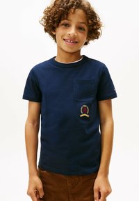 Camiseta de algodón azul marino de manga corta, con un bolsillo en el pecho izquierdo y un escudo bordado en rojo y dorado en la parte inferior.