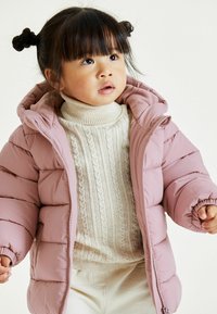 Veste rose bouffante avec une capuche, présentant des sections matelassées horizontales et des poignets élastiques, portée par-dessus un pull à col roulé en maille torsadée crème.